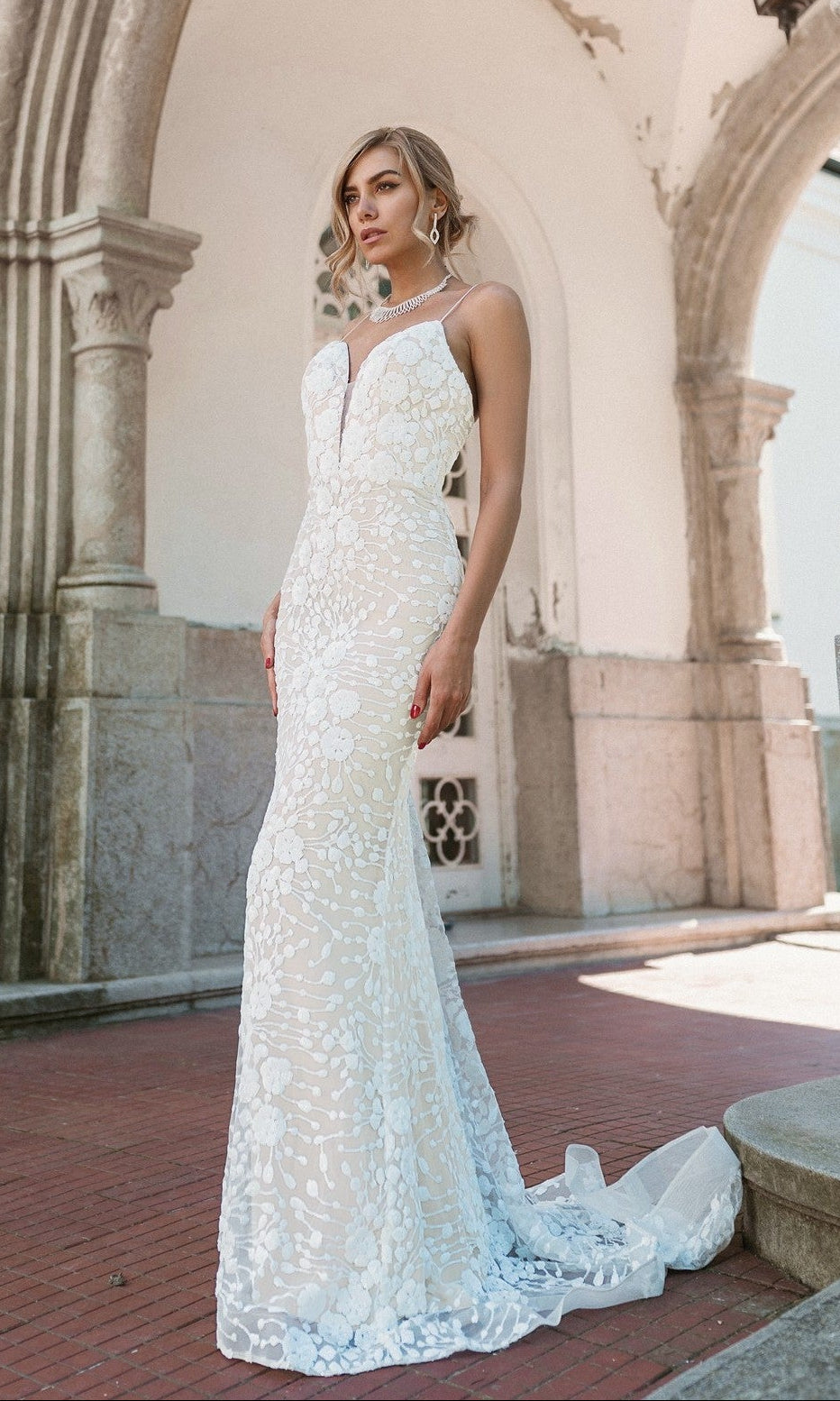 A woman wears the Tina Holly BB008 Long White Lace Formal Dress in white/ivory, standing outside under an arched stone structure and facing forward with a neutral expression.